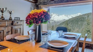 Een eetruimte met een tafel gedekt voor vier personen, een vaas met bloemen, trofeeën op een kast, en een uitzicht op een berg door een groot raam.
