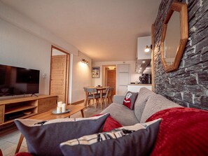 A modern living room with a flat-screen TV, wooden furniture, a stone wall, and a kitchen area in the background. - Superb apartment in station center - 6 guests (La Plagne)