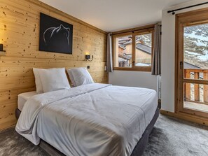 A bedroom with a wooden headboard, a large bed with white bedding, a mounted picture, and a window with a view of buildings and trees. - Comfortable apartment, 6 guests, Belle Plagne (Belle Plagne)