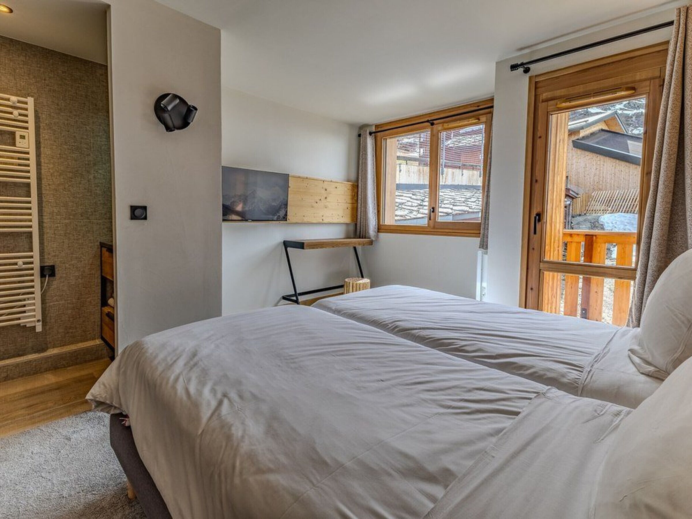 Ein Schlafzimmer mit zwei Betten, einem Holz-Kopfteil, einem an der Wand befestigten Fernseher und einem Fenster mit Blick auf eine Holzterrasse.