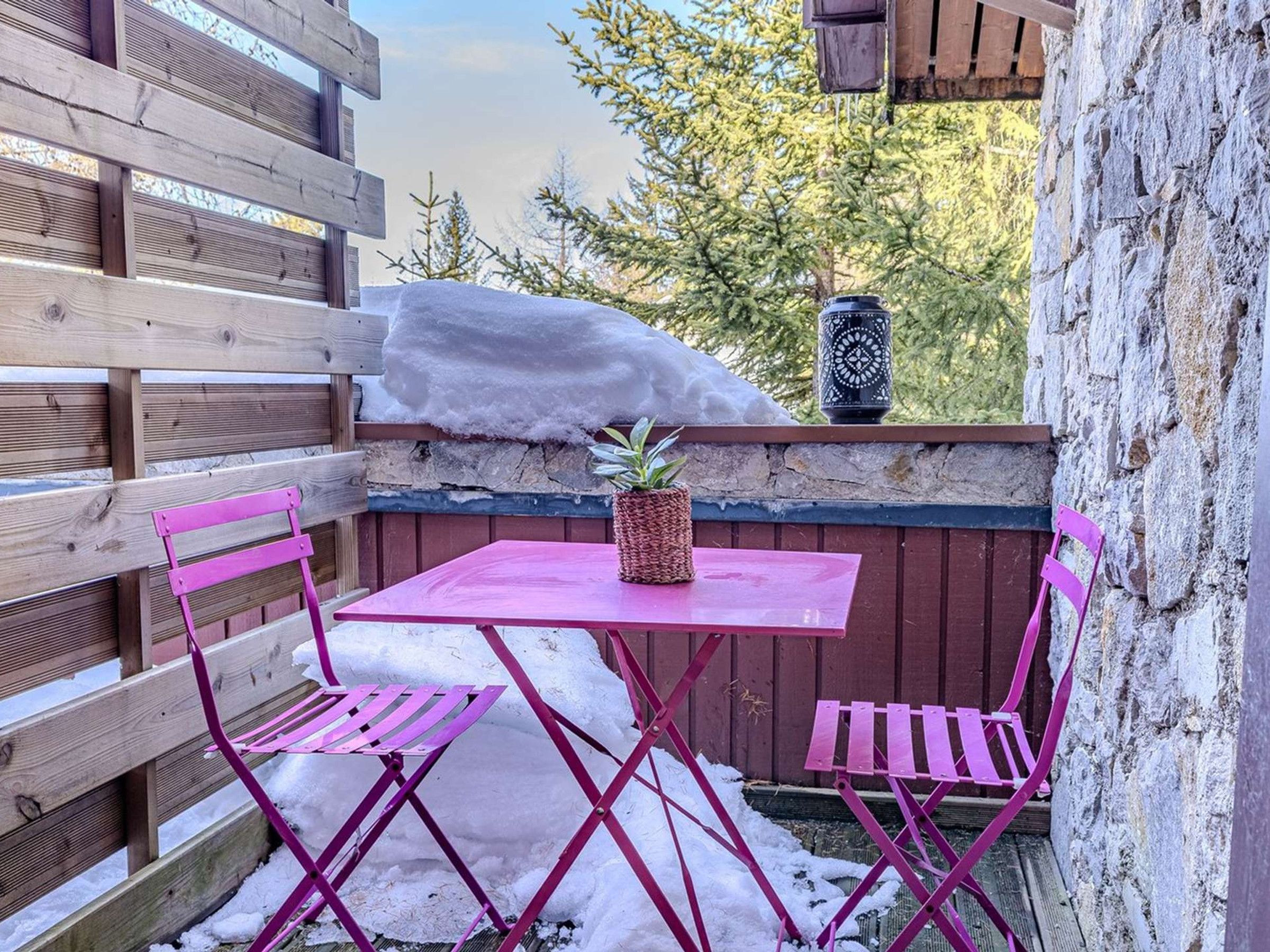 Een houten terras met een roze tafel en stoelen, een bloempot en een stenen muur.