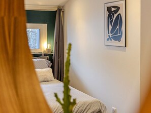 Ein Schlafzimmer mit Bett, Nachttisch, Lampe, Fenster und einem an der Wand hängenden Kunstwerk.