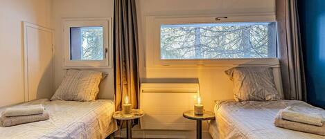 A bedroom with two beds, a window, a bedside table with a lamp, and a curtain.