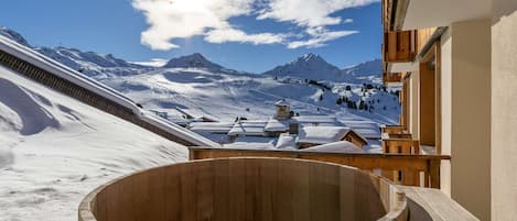 A wooden hot tub on a balcony overlooking a snowy mountain landscape.