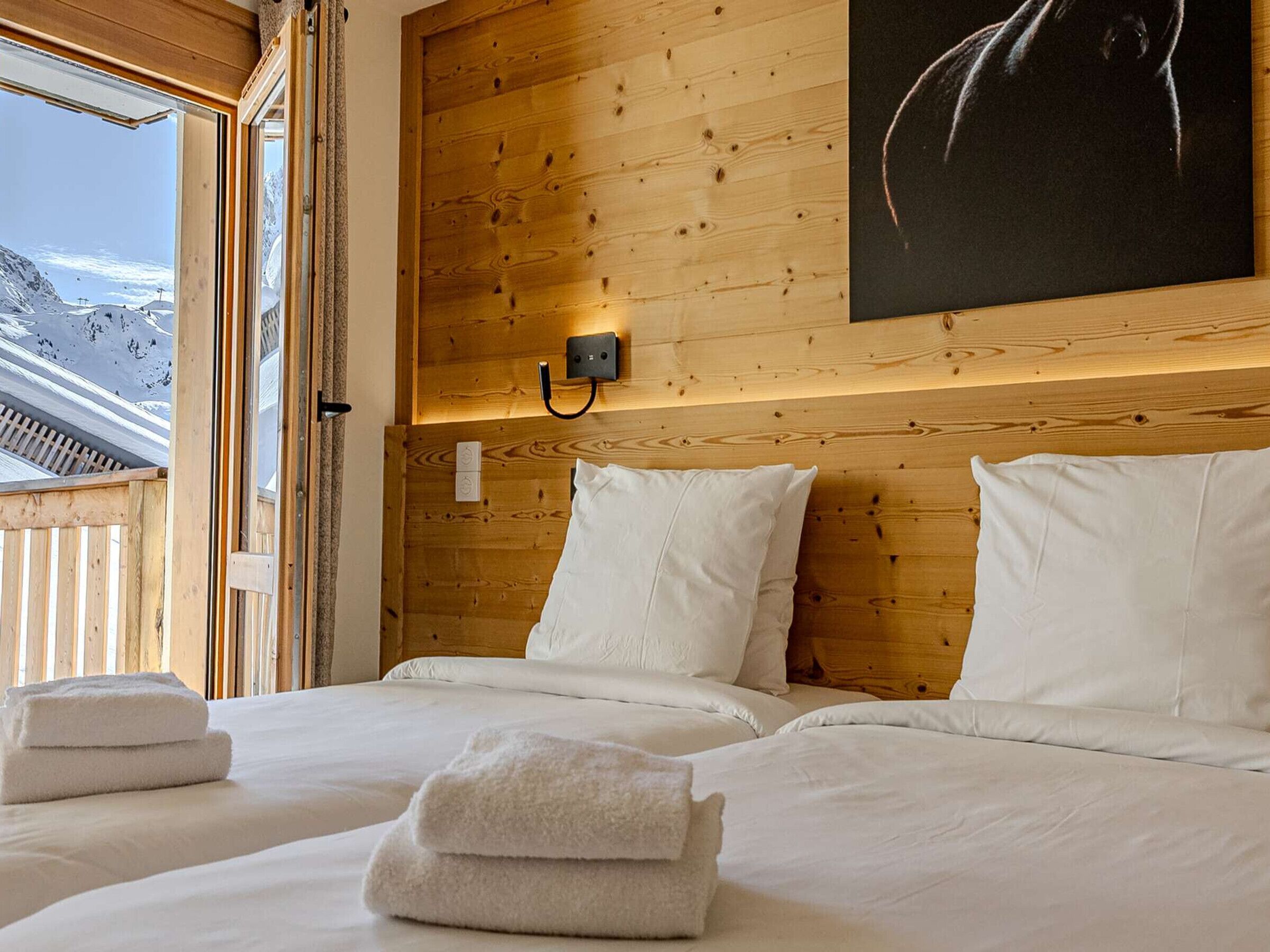 Une chambre avec une tête de lit en bois, un lit recouvert de draps blancs, un tableau accroché au mur et une fenêtre donnant sur les montagnes.