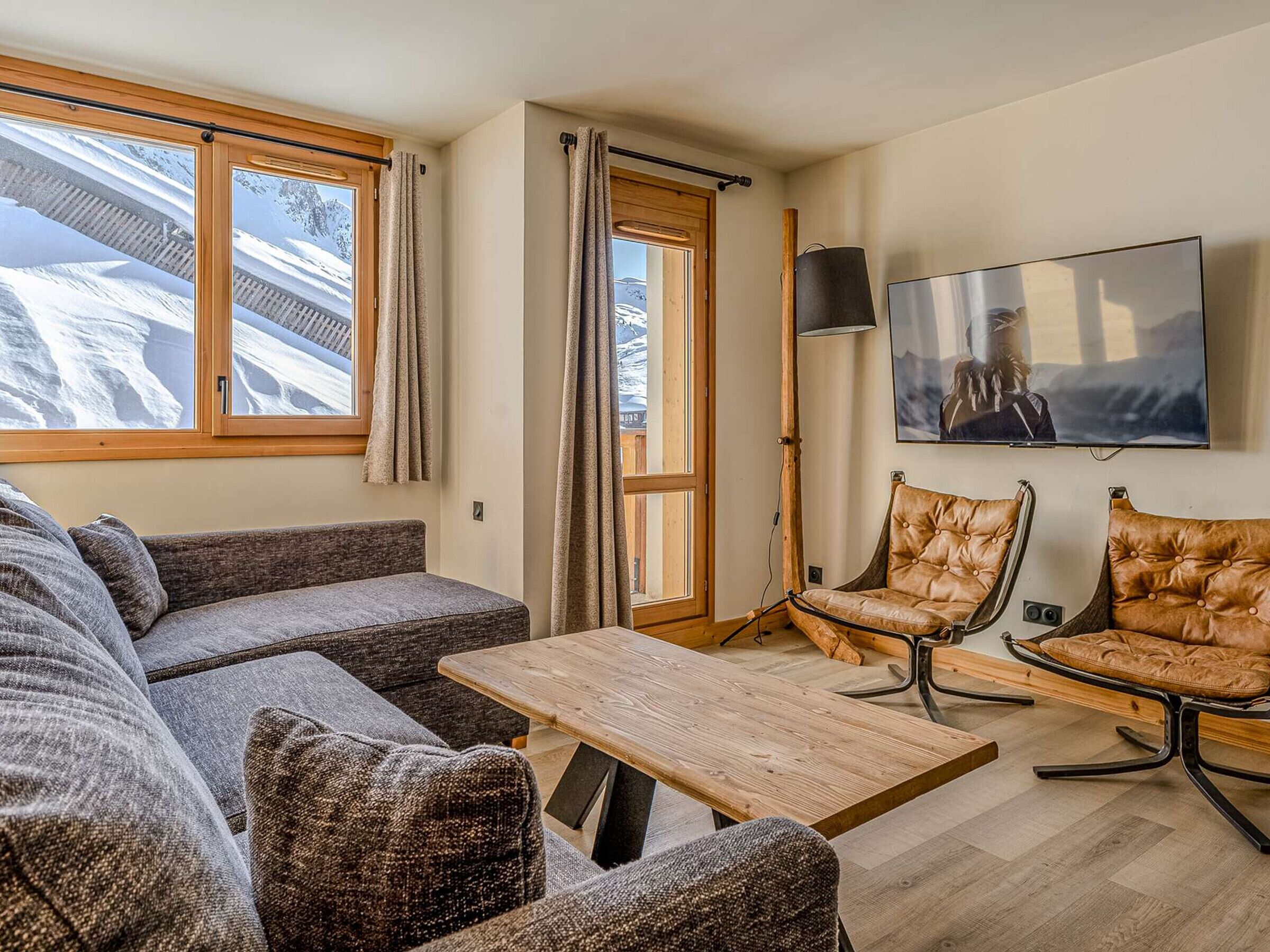 Ein modernes Wohnzimmer mit einer Couch, zwei Ledersesseln, einem Holztisch, einem Fernseher und großen Fenstern mit Vorhängen.