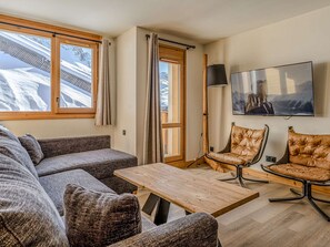 Ein modernes Wohnzimmer mit einer Couch, zwei Ledersesseln, einem Holztisch, einem Fernseher und großen Fenstern mit Vorhängen.