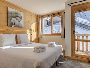 Ein Schlafzimmer mit einem Bett, Kissen, einer Wandleuchte, einem Fenster mit Blick auf eine verschneite Landschaft und einer Holztür.