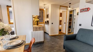 Ein modernes Wohnzimmer mit einer Couch, einem Essbereich mit Tisch für zwei Personen und einer Küche im Hintergrund.