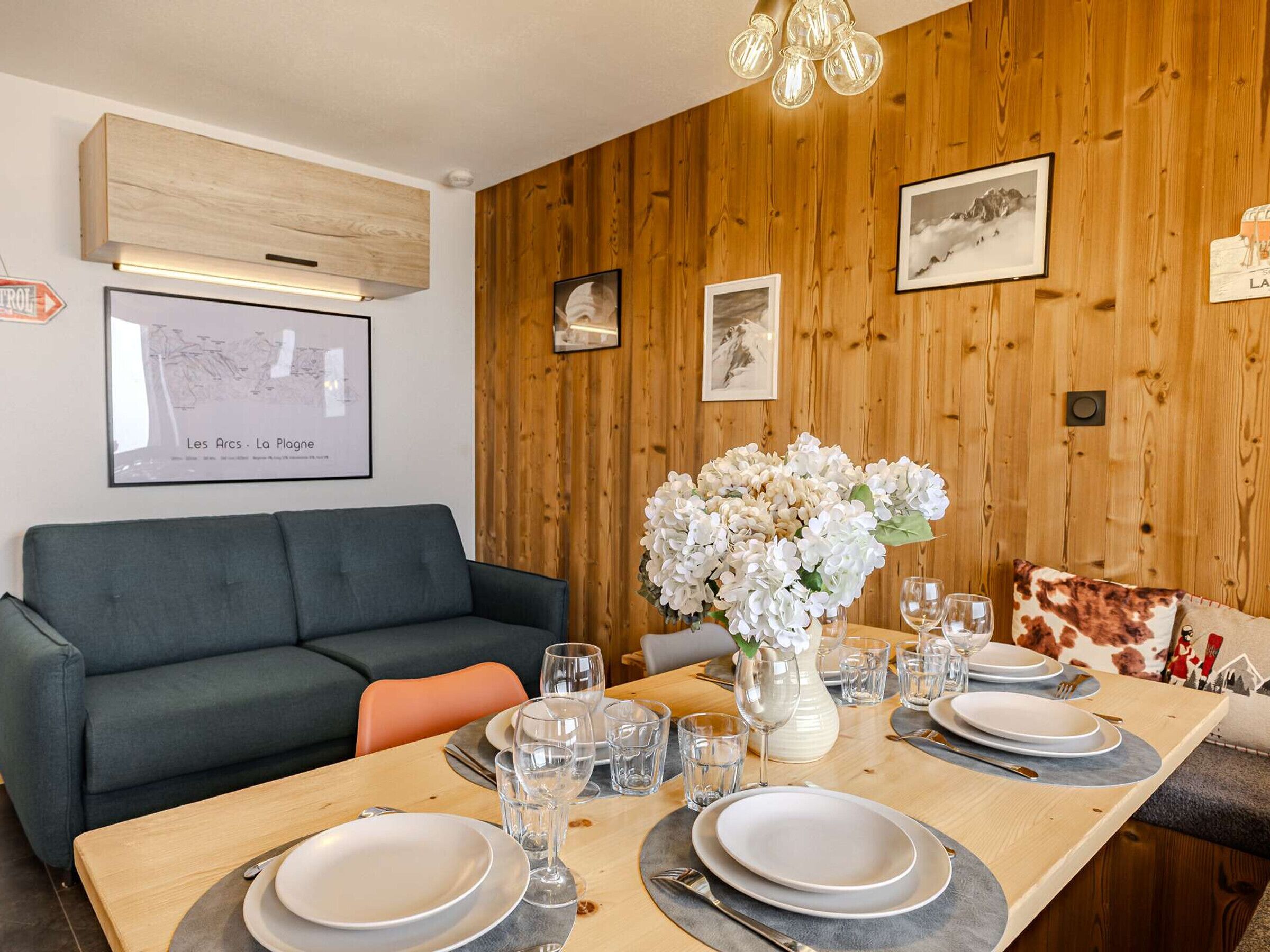 A dining area with a wooden table set for four, a sofa, and framed pictures on the wall.