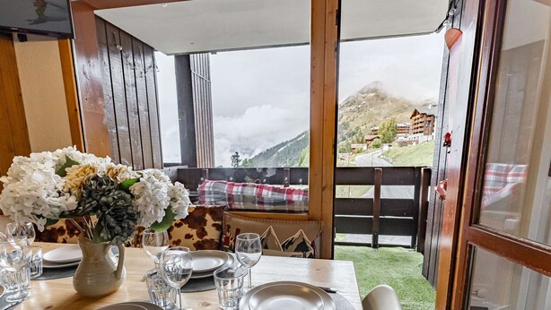 Ein Esstisch ist gedeckt mit Tellern, Gläsern und Besteck, eine Blumenvase steht darauf, und durch große Fenster bietet sich der Blick auf eine Berglandschaft.