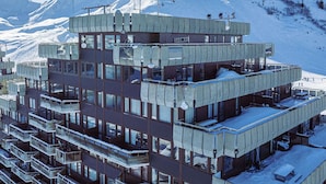 Ein mehrstöckiges Gebäude mit Balkonen, im Hintergrund eine Skipiste und eine gebirgige Landschaft.