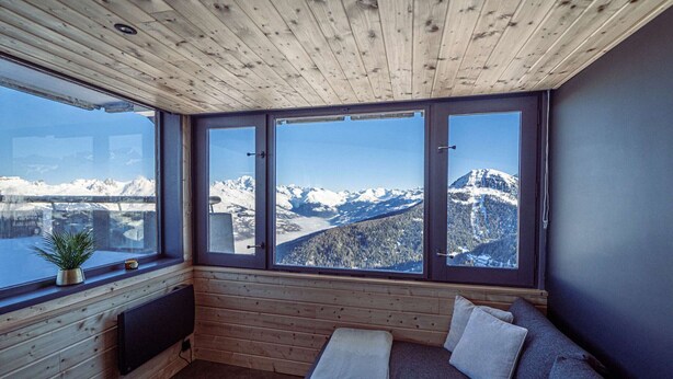 Eine Holzhütte mit einer Couch, einem kleinen Tisch und Blick auf schneebedeckte Berge.