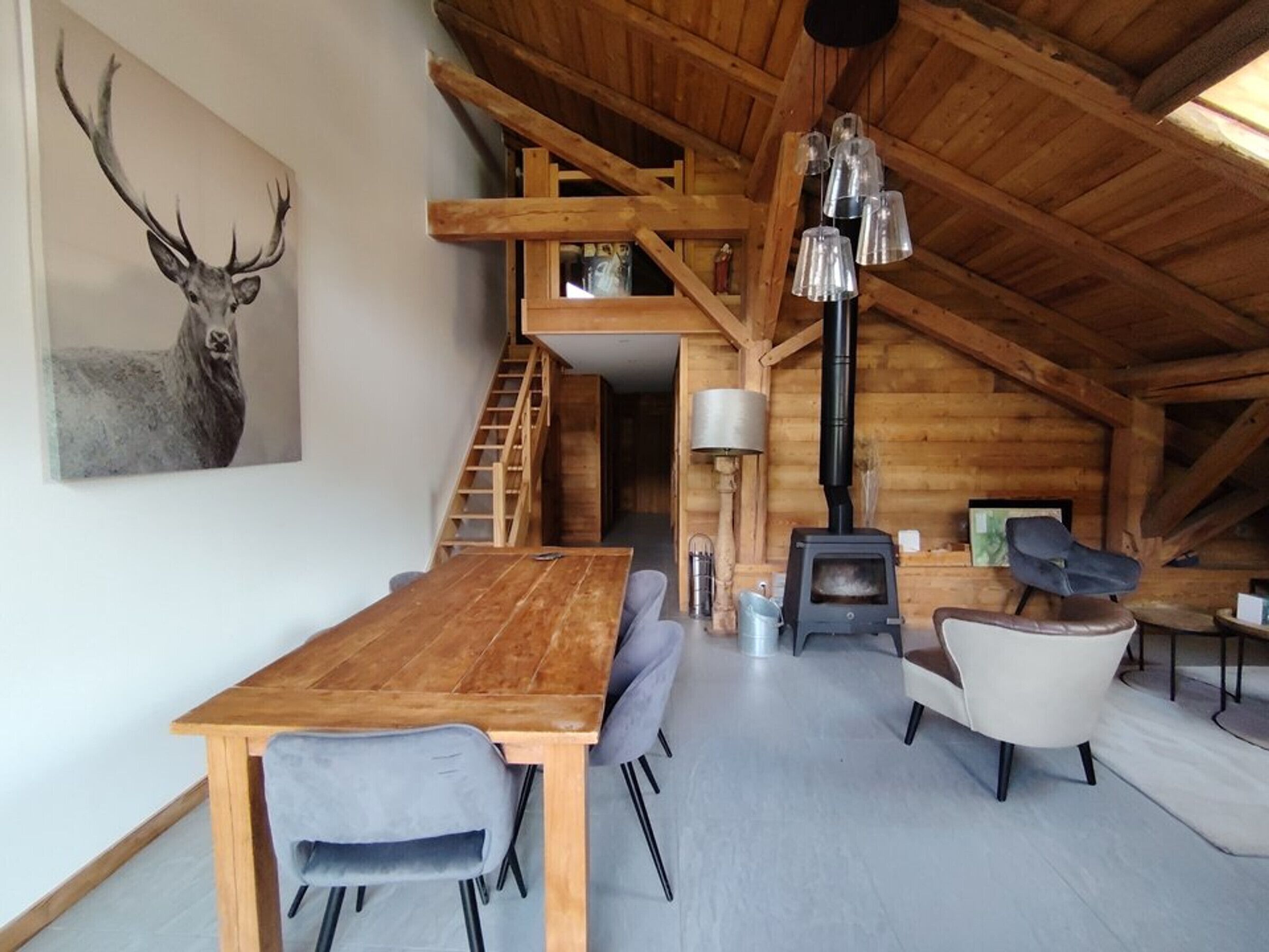 Een houten interieur met een eettafel, stoelen, een trap, een open haard en een schilderij van een hert.