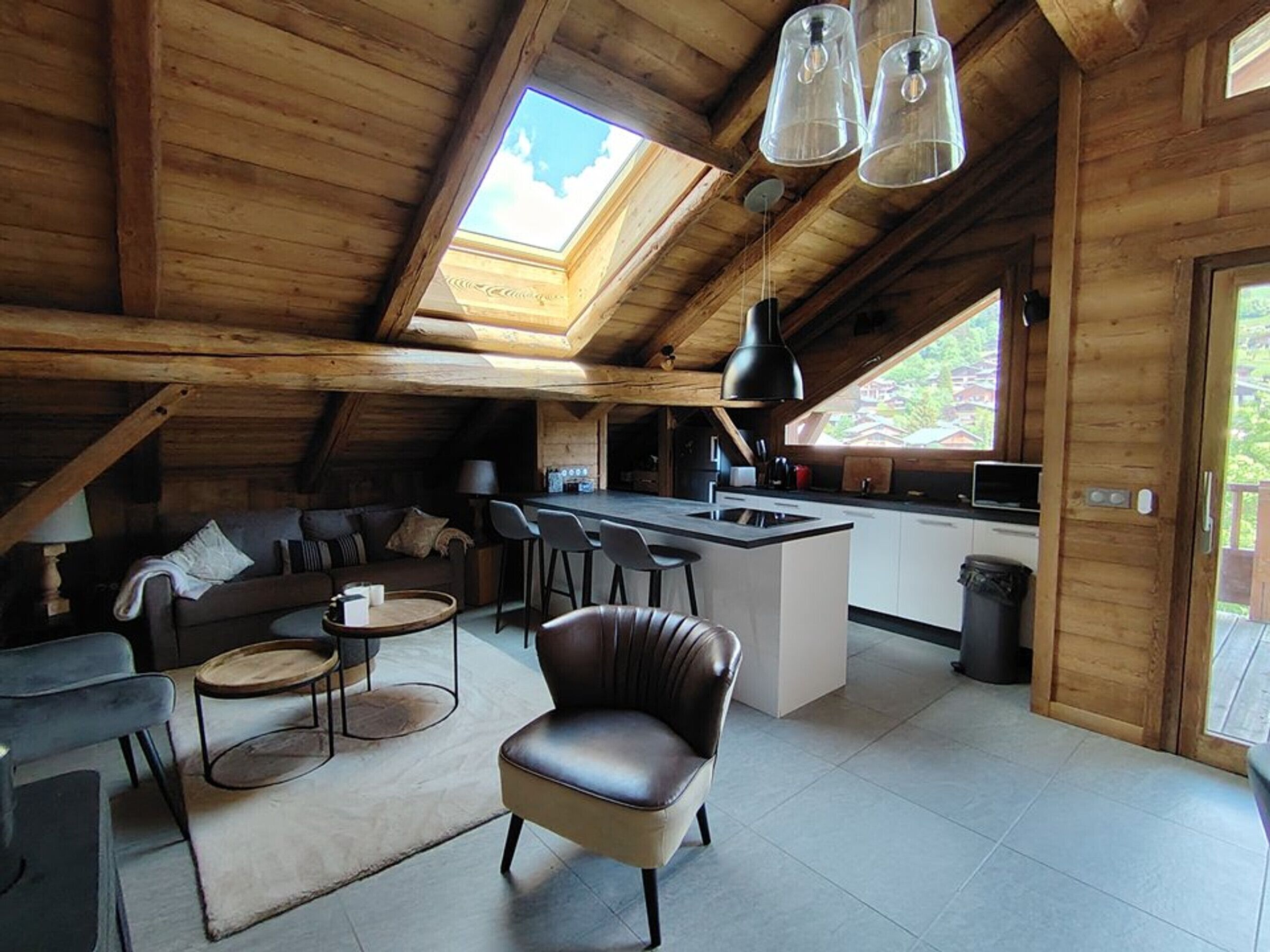 A spacious loft with a high ceiling, wooden beams, a cozy seating area with a sofa and chairs, a kitchen with a bar, and a skylight.
