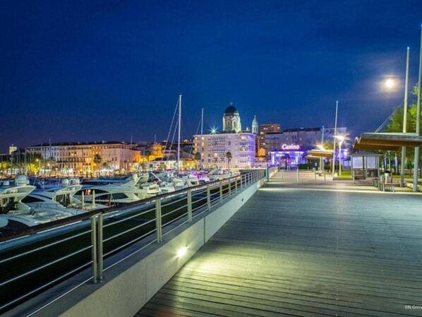 Eine Marina bei Nacht mit vertäuten Booten, ein Casino im Hintergrund und eine Promenade, die zu einem überdachten Bereich führt.