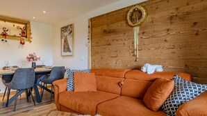 A modern living room with a wooden accent wall, a sofa with patterned cushions, a dining area with chairs, and a decorative wall hanging.
