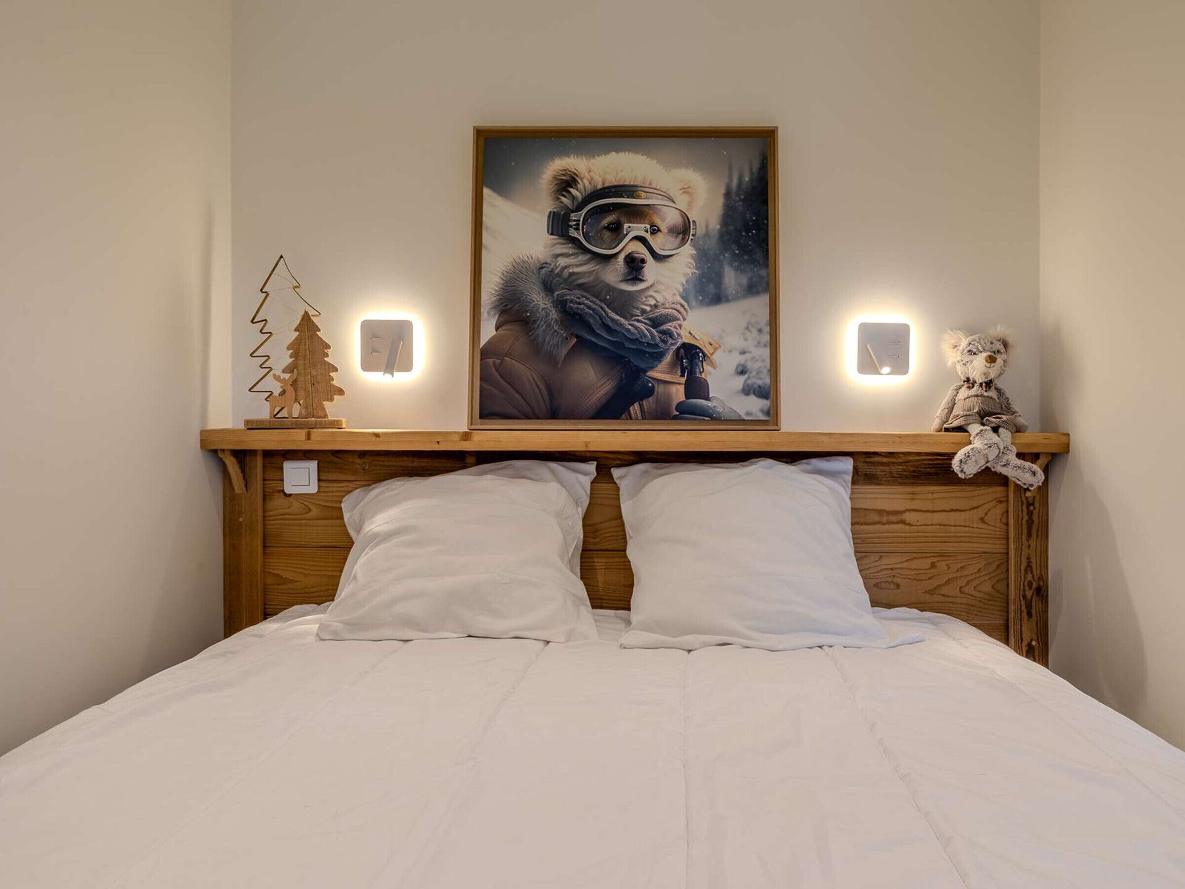 A bed with white bedding, a wooden headboard, a framed picture of a bear in winter gear, a small wooden Christmas tree, and two wall-mounted lights.