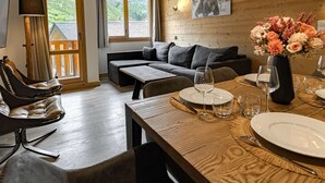 Ein gemütliches Wohnzimmer mit einer Couch, einem Esstisch für zwei Personen und Blick ins Freie durch große Fenster.