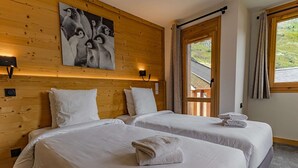 Ein Hotelzimmer mit zwei Betten, Holzwänden, einem Gemälde von Pinguinen, einem Fenster mit Vorhängen und Blick ins Grüne.