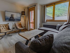 Ein modernes Wohnzimmer mit einer Couch, Sesseln, einem Tisch, einem Fernseher und einem Fenster mit Blick auf eine Berglandschaft.