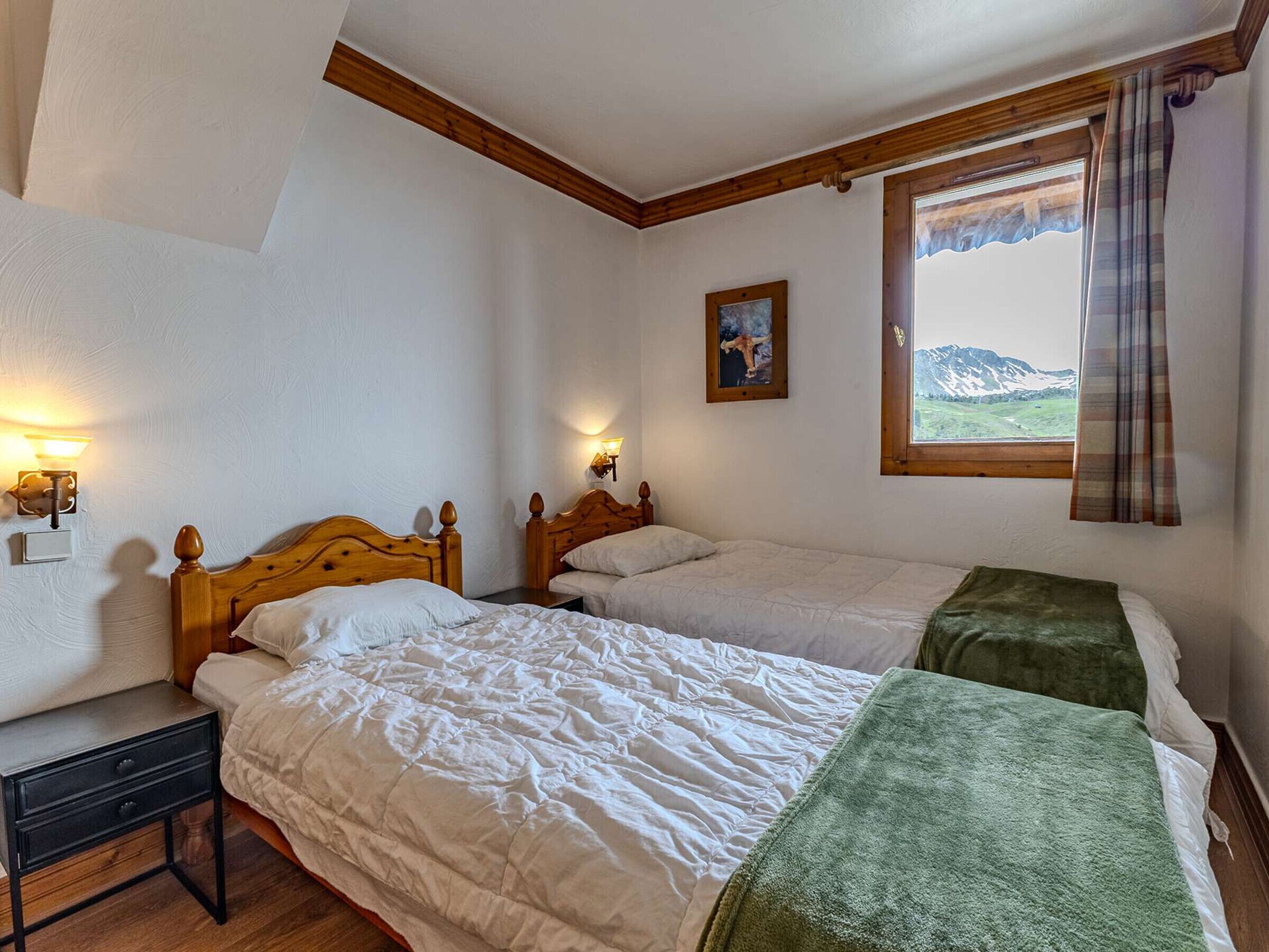 Ein Zimmer mit zwei Holzbett, einem Nachttisch, einer Wandleuchte, einem Fenster mit Bergblick und einem Bild an der Wand.