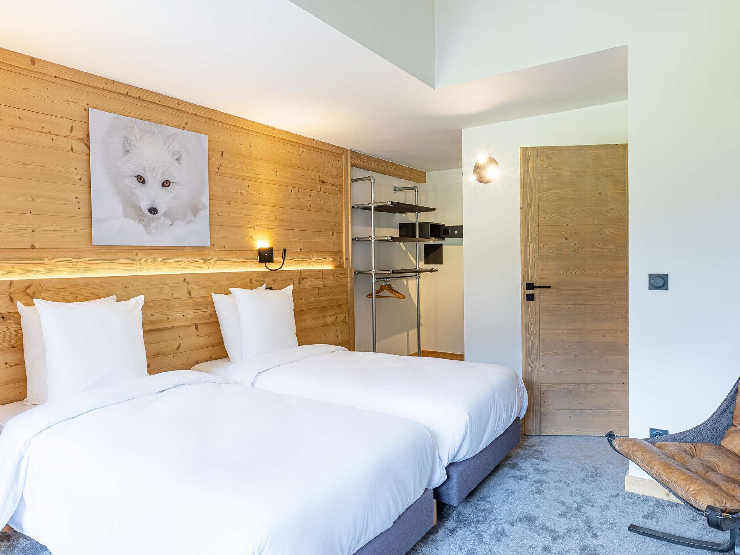 Ein Schlafzimmer mit Holz-Kopfteil, einem Bett mit weißer Bettwäsche, einer Wandleuchte, einem Kleiderständer und einer Tür.