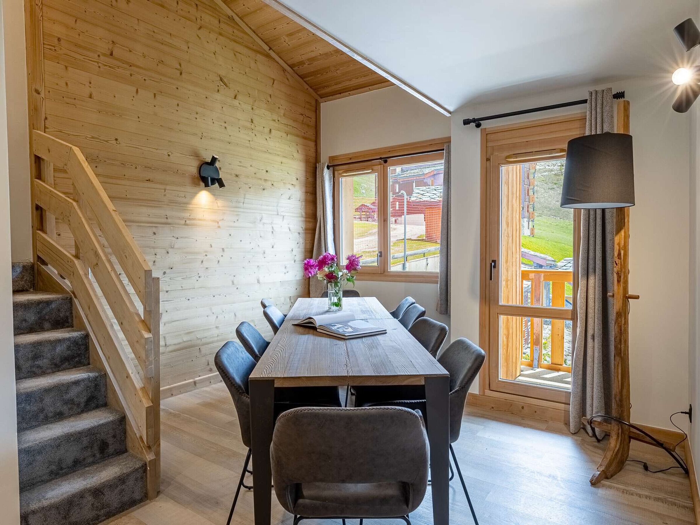 A dining area with a wooden table and chairs, a vase with flowers, a staircase, and a window with a view of the outside.