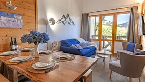 Una mesa de comedor de madera dispuesta con platos y vasos, un sofá azul, un sillón gris y un ventanal con vistas a la montaña.