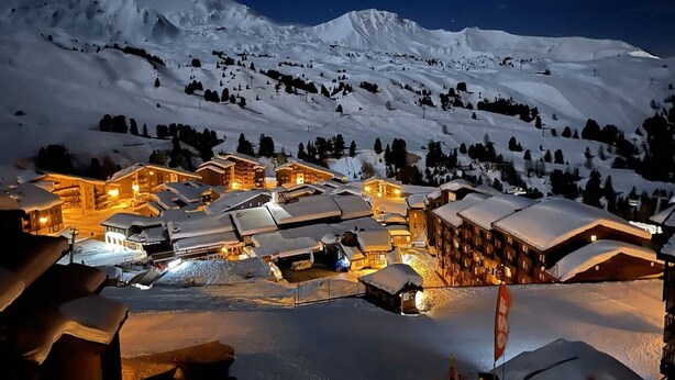 Un complejo turístico de montaña cubierto de nieve de noche, con edificios iluminados y una vista despejada del paisaje nevado circundante.