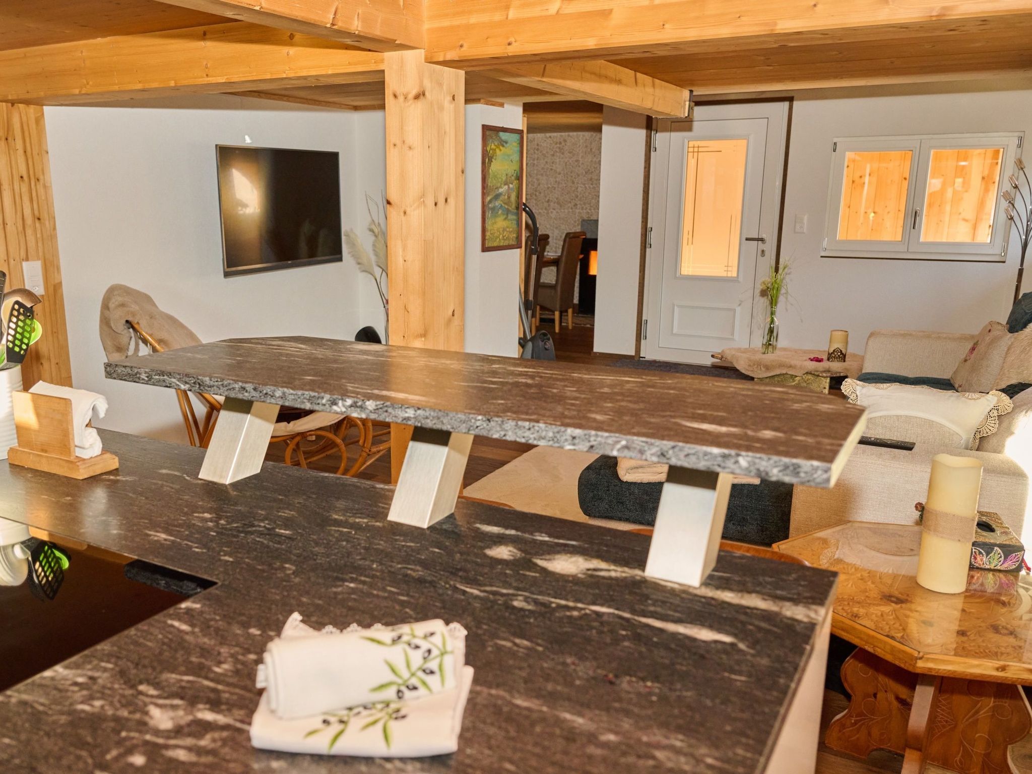 A modern kitchen with a granite countertop, wooden beams, a dining area with a sofa, and a TV mounted on the wall.