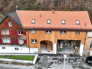 Une maison en bois à deux étages, avec un toit recouvert de tuiles rouges, un garage doté d’un escalier en bois et un petit jardin.