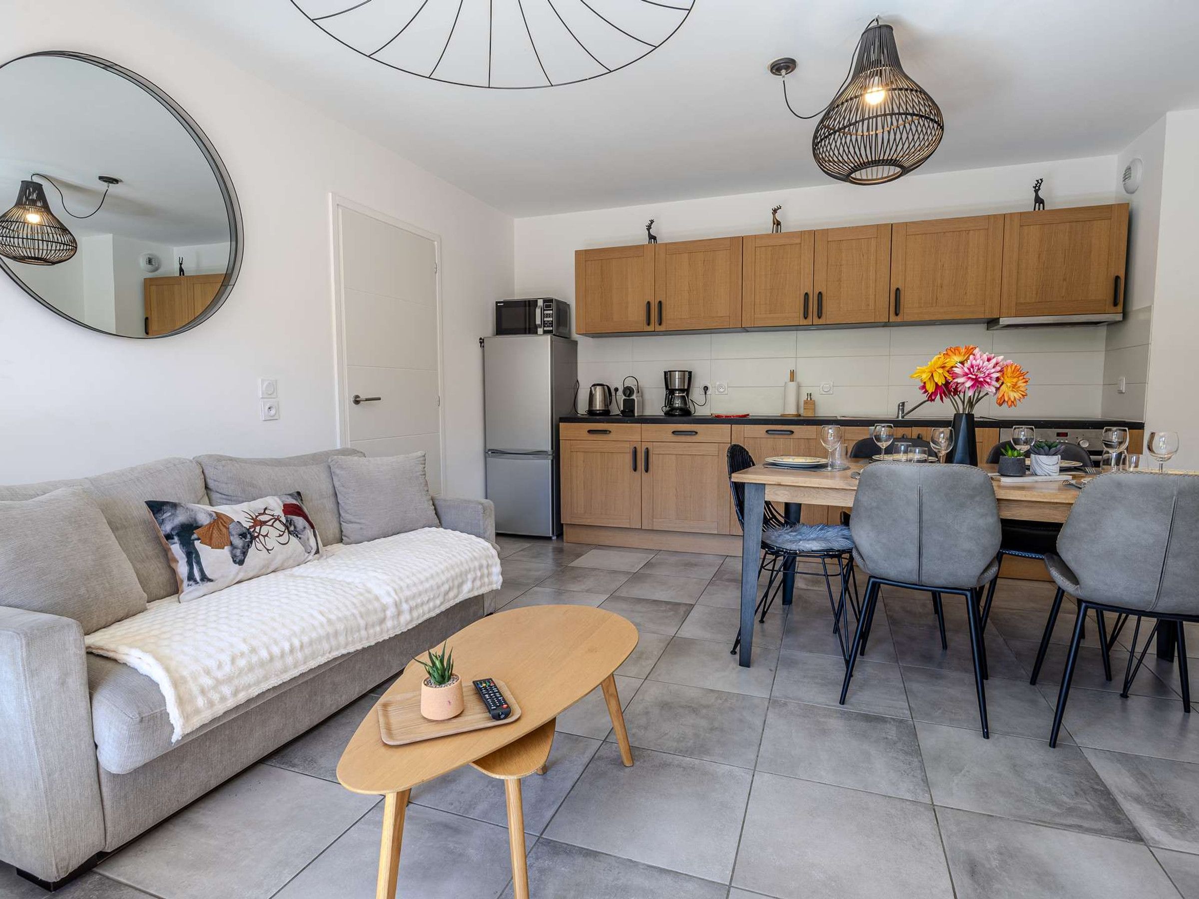 Een gezellig, compact woongedeelte met een bank, een salontafel, een eethoek met stoelen, een keuken met houten kastjes en een hangende lamp.