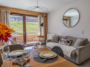 A dining area with a table set for two, a sofa, a mirror, and a view of greenery outside. - Luxury apartment, prime location - 6 guests (La Plagne)