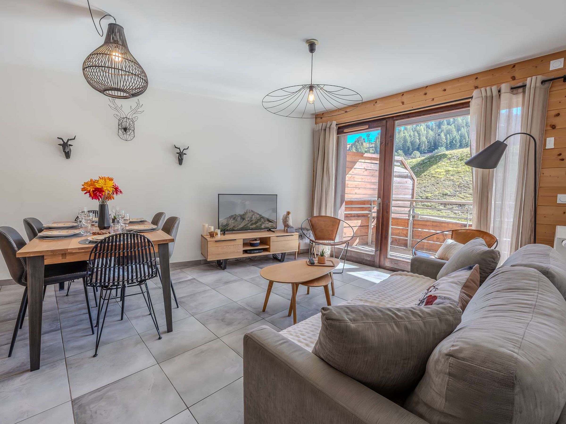 A modern living room with a sofa, dining table, and chairs, a TV, and a balcony with a view of the outdoors.