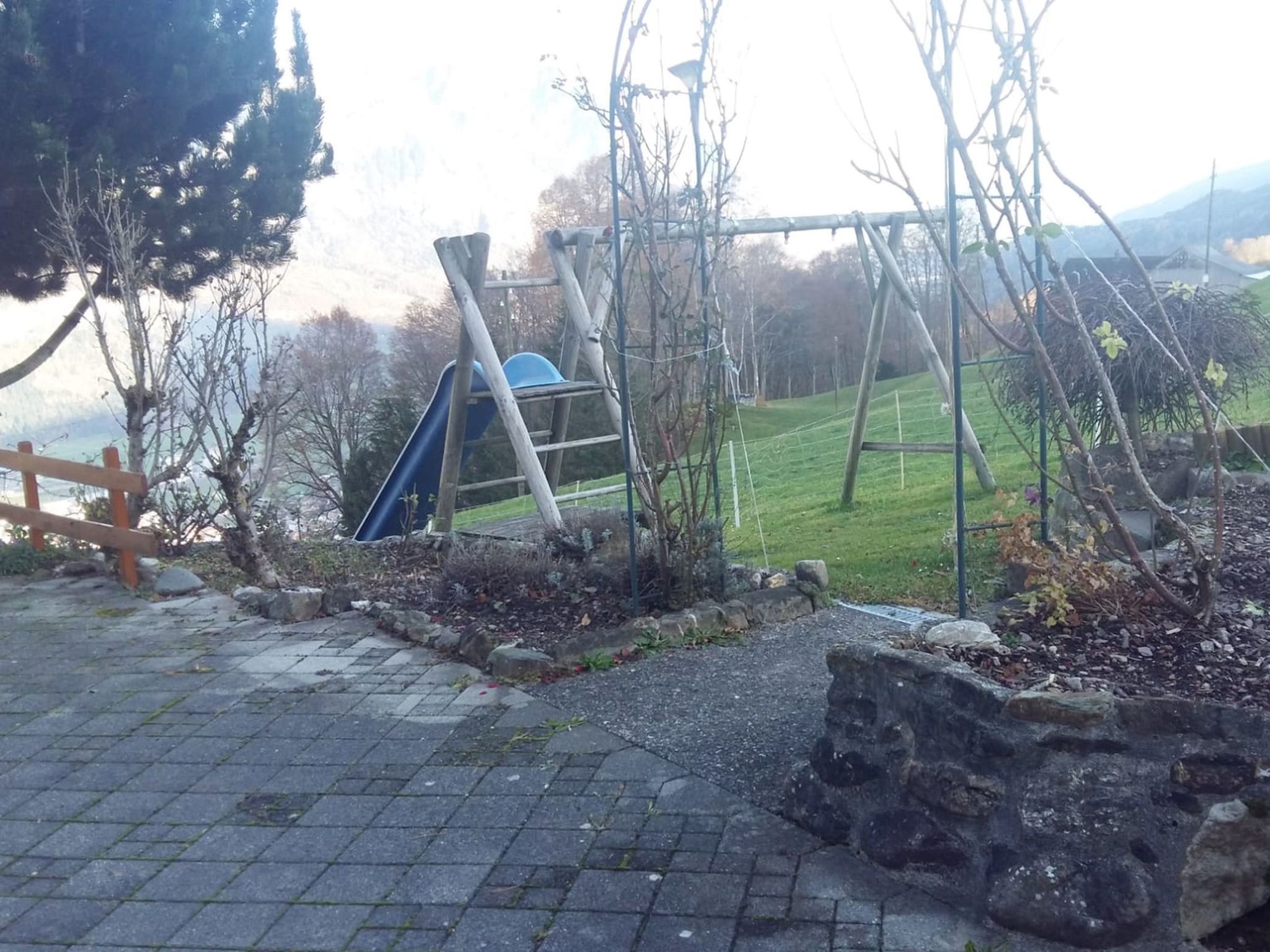 Ein Spielplatz mit Rutsche und Schaukeln, umgeben von einer Steinmauer und Bäumen.