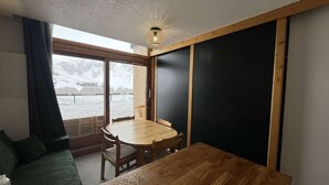 A room with a wooden dining table, a green sofa, a large window with a view of a snowy landscape, and a ceiling light fixture. - Modern studio at the foot of the slopes - 6 guests (Plagne Bellecôte)