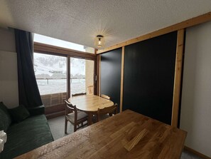 Ein Zimmer mit einem Holzes Esstisch, einem grünen Sofa, einem großen Fenster mit Blick auf eine verschneite Landschaft und einer Deckenleuchte.