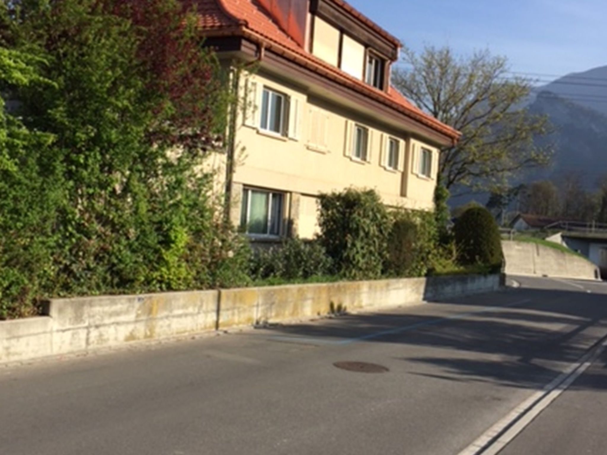 Un bâtiment à deux étages, avec un toit recouvert de tuiles rouges, un mur de soutènement en pierre et un ciel dégagé.