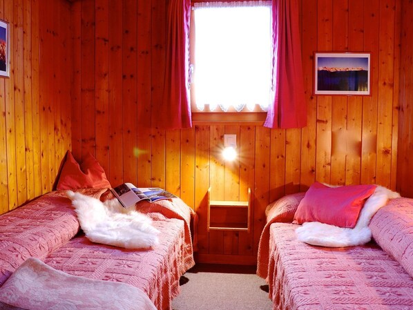 A wooden cabin room with two beds, a window with curtains, and a small table.