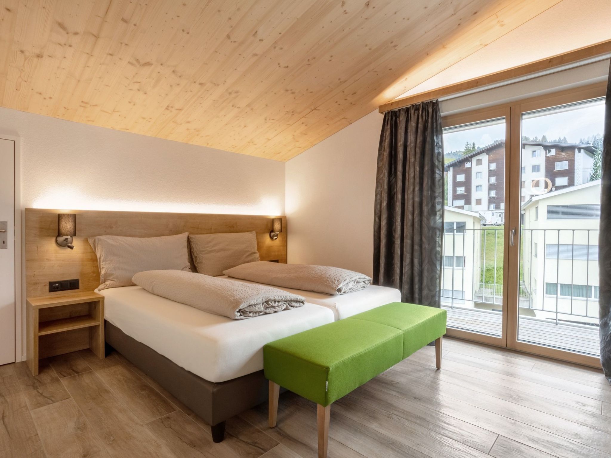 A bedroom with a bed, bedside tables, a bench, and a view of buildings through a large window.