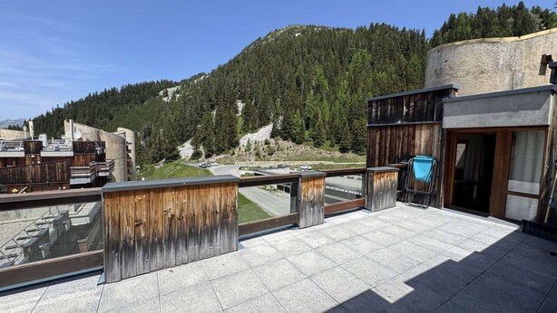 Una terraza en la azotea con vistas a una montaña arbolada, una zona pavimentada y un edificio con fachada de madera.