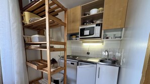 Cocina compacta con armarios blancos, microondas, lavamanos y estantería de madera.