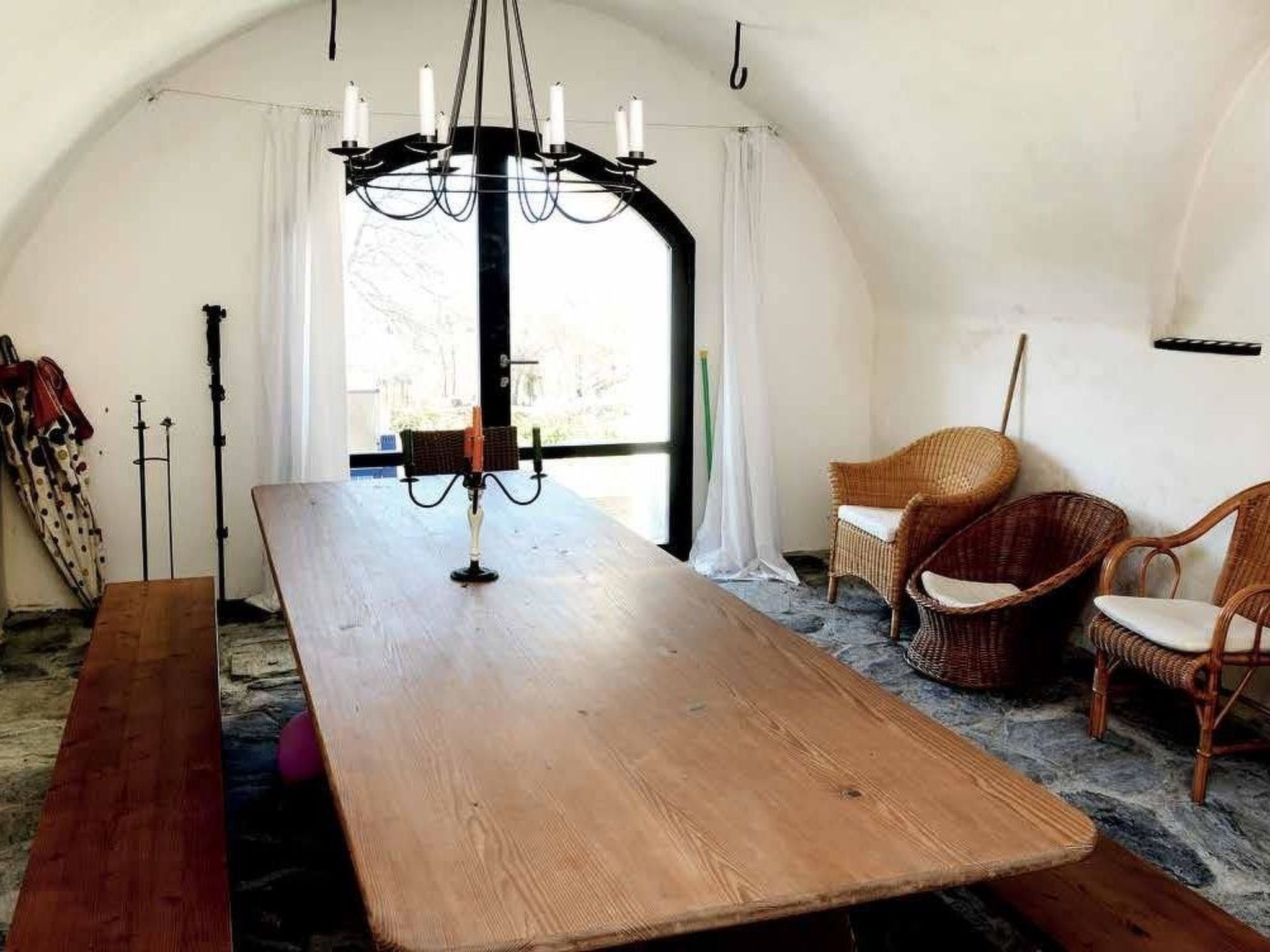 A room with a large wooden table, two wicker chairs, a chandelier, and a window with curtains.