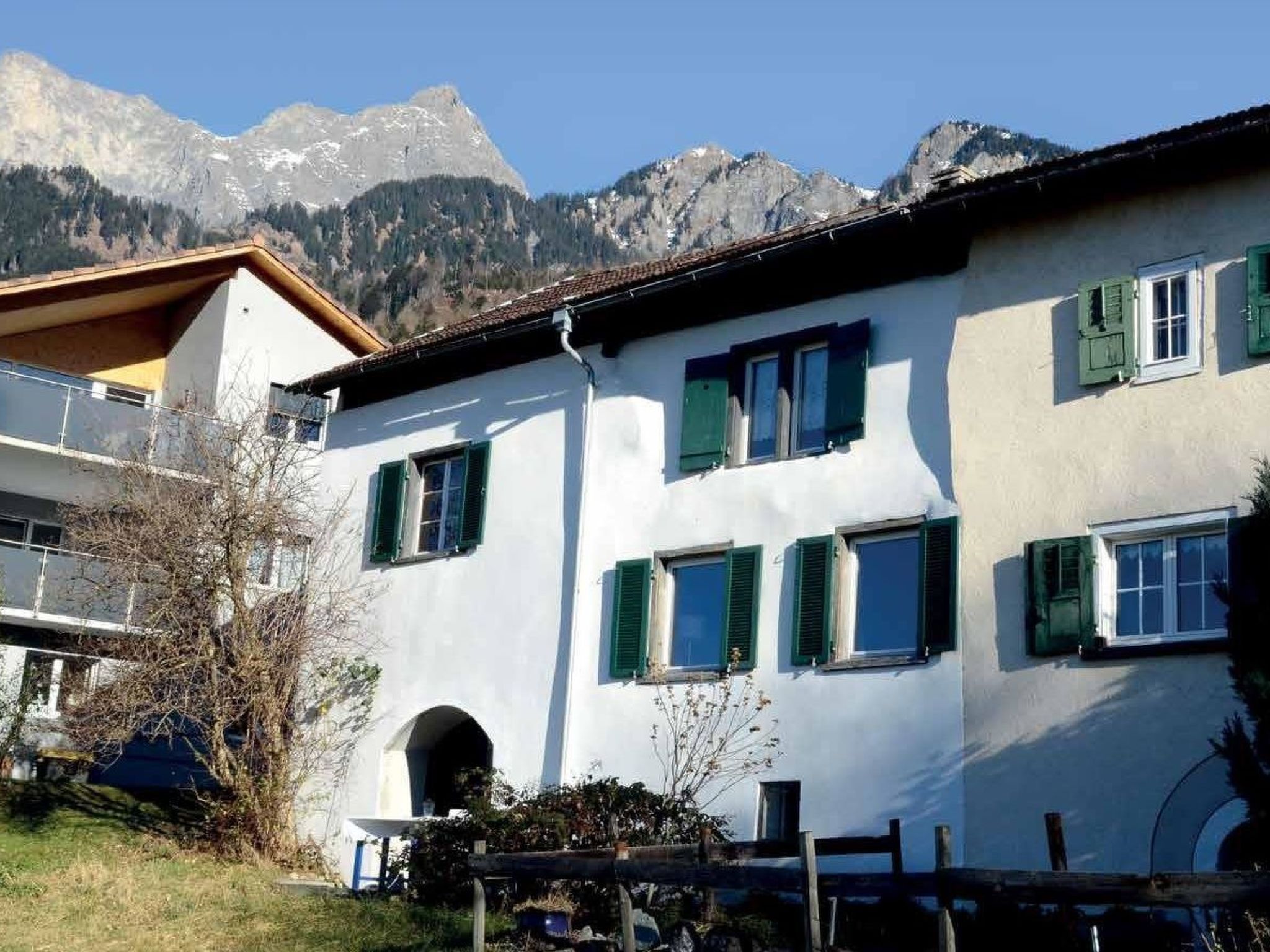 A white building with green shutters, a balcony, and a mountain backdrop.