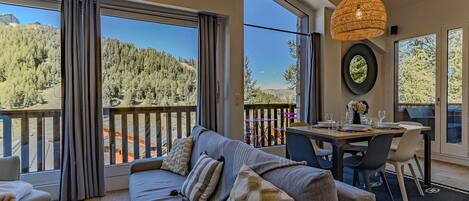 A modern living room with a sofa, dining table, and chairs, large windows with a mountain view, and a wooden ceiling.