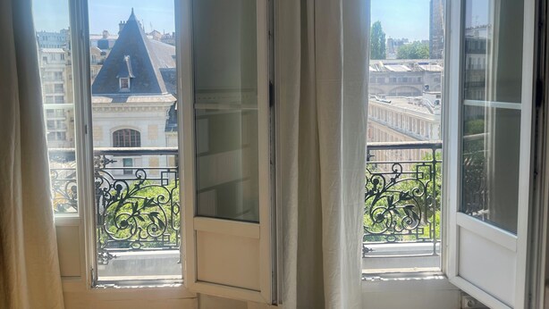 Ein Fenster mit Blick auf ein Gebäude mit spitzer Dachform und Balkonen mit aufwendigen Eisenengelgittern.