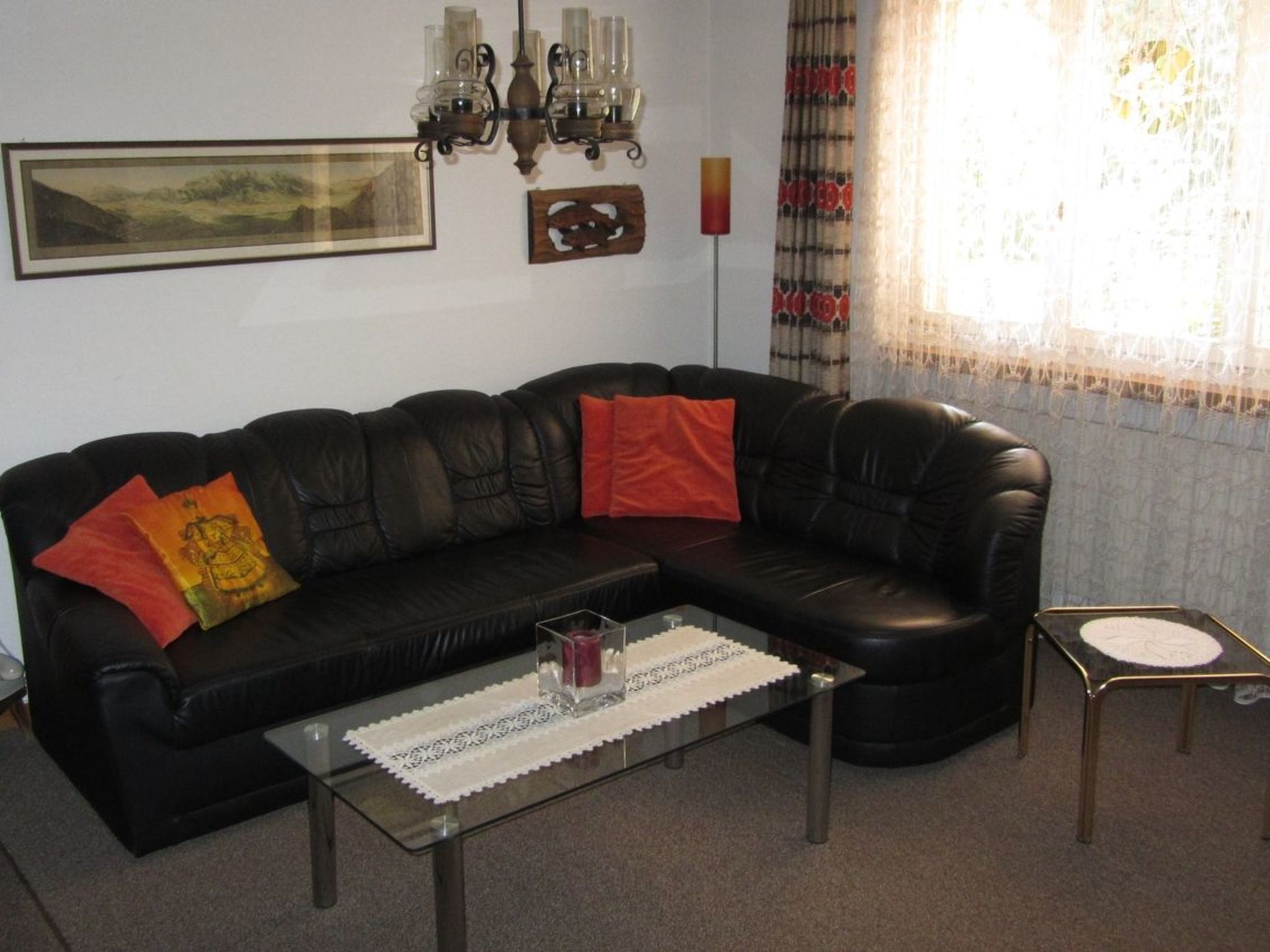 A black leather sofa with red and yellow cushions, a glass coffee table with a lace doily, a candle, and a small side table with a clock.
