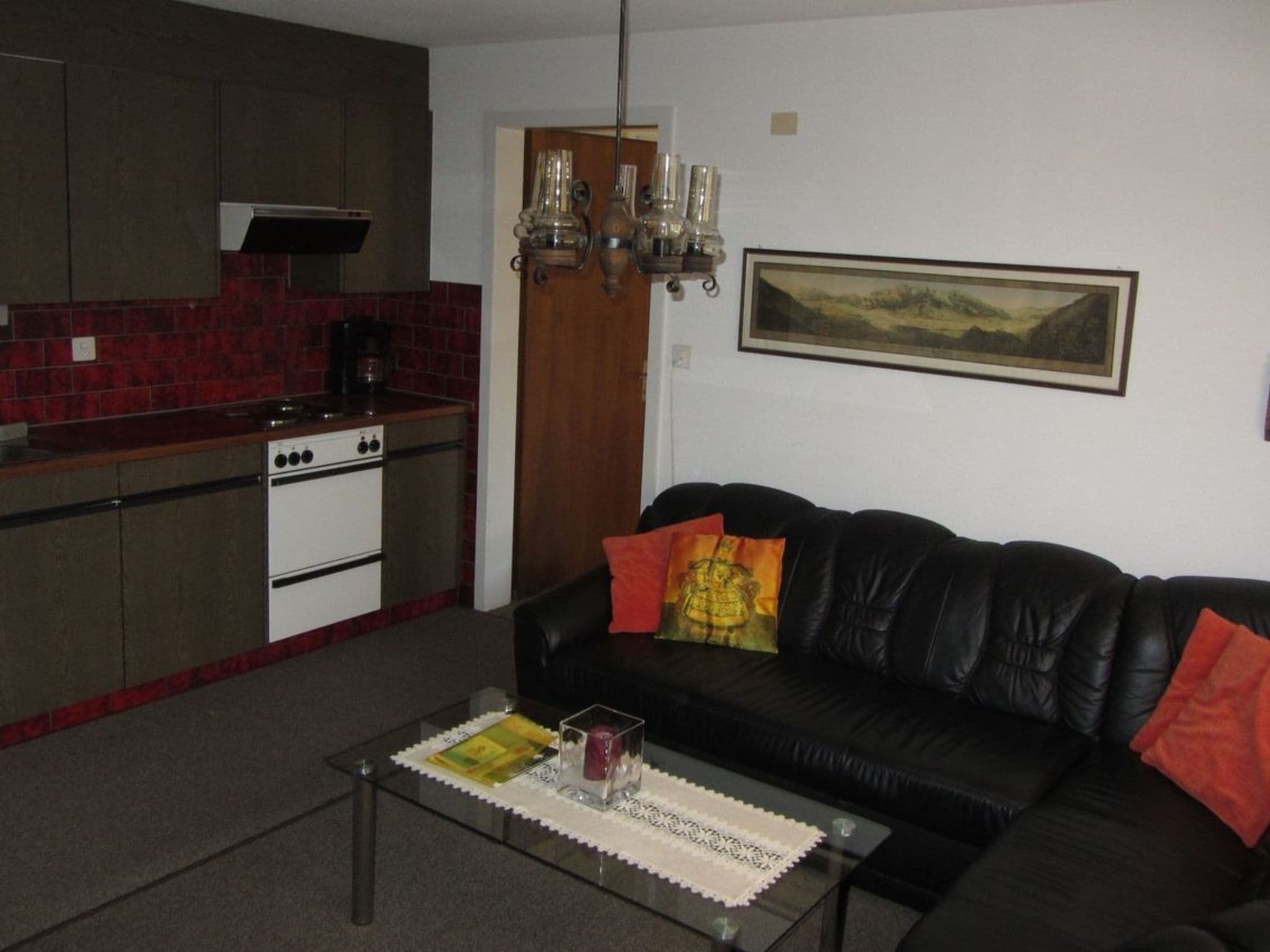 A living room with a black leather sofa, a glass coffee table, and a kitchen area with red tiles and a stove.