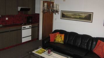 A living room with a black leather sofa, a glass coffee table, and a kitchen area with red tiles and a stove.